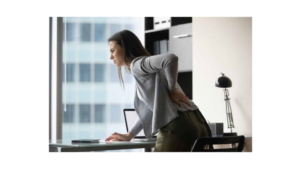 Woman bent over in pain on desk bracing her lower back sciatic pain.
